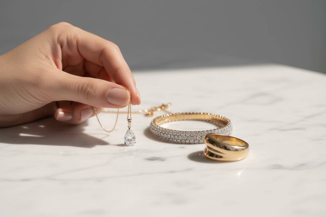Fine jewelry pieces arranged on white marble — diamond pendant necklace, pavé bracelet, and gold ring — with warm studio lighting and an elegant hand examining quality.