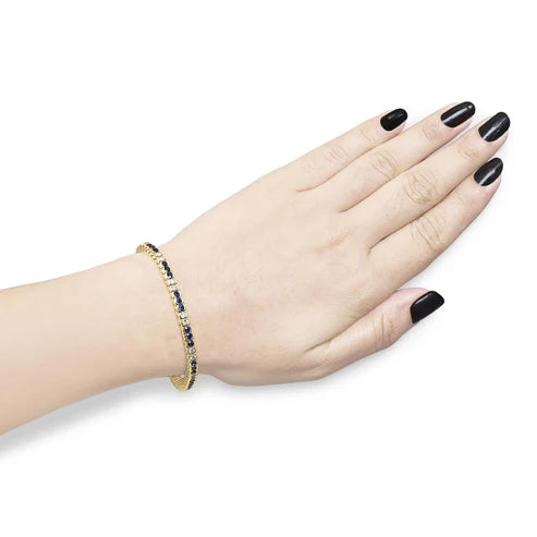 Load video: Video demonstration of a 14K yellow gold 1 ¾ cttw diamond and 5 cttw blue sapphire bezel‑set tennis bracelet on a model’s wrist.