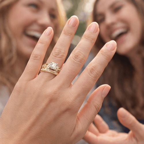 Close-up of a hand wearing a gold ring with a diamond, with two people in the background.